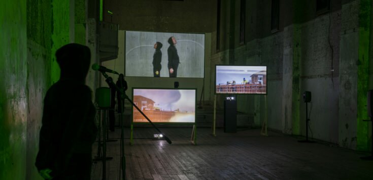 A person stands watching a set of three projected screens, in a darkened gallery space. There is a slight green light on the concrete walls.